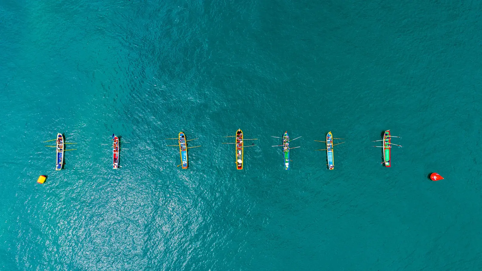 Sete canoas de regata alinhadas sobre a água turquesa, vistas de cima