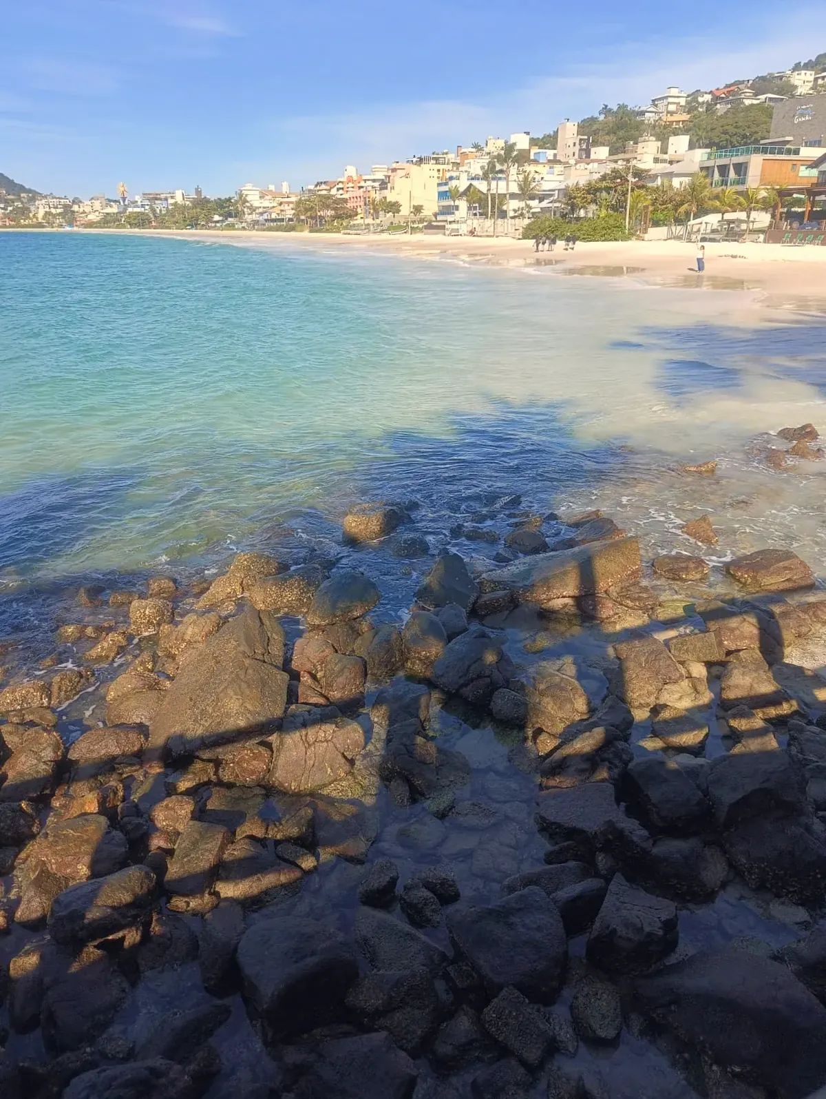 Água clara sobre as pedras, com a praia e as casas do morro ao fundo